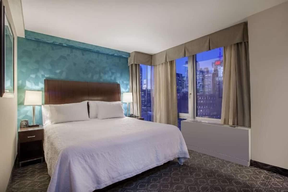 Une chambre d’hôtel avec un grand lit, une table de chevet, une lampe, une fenêtre avec des rideaux et une vue sur le paysage urbain au crépuscule.