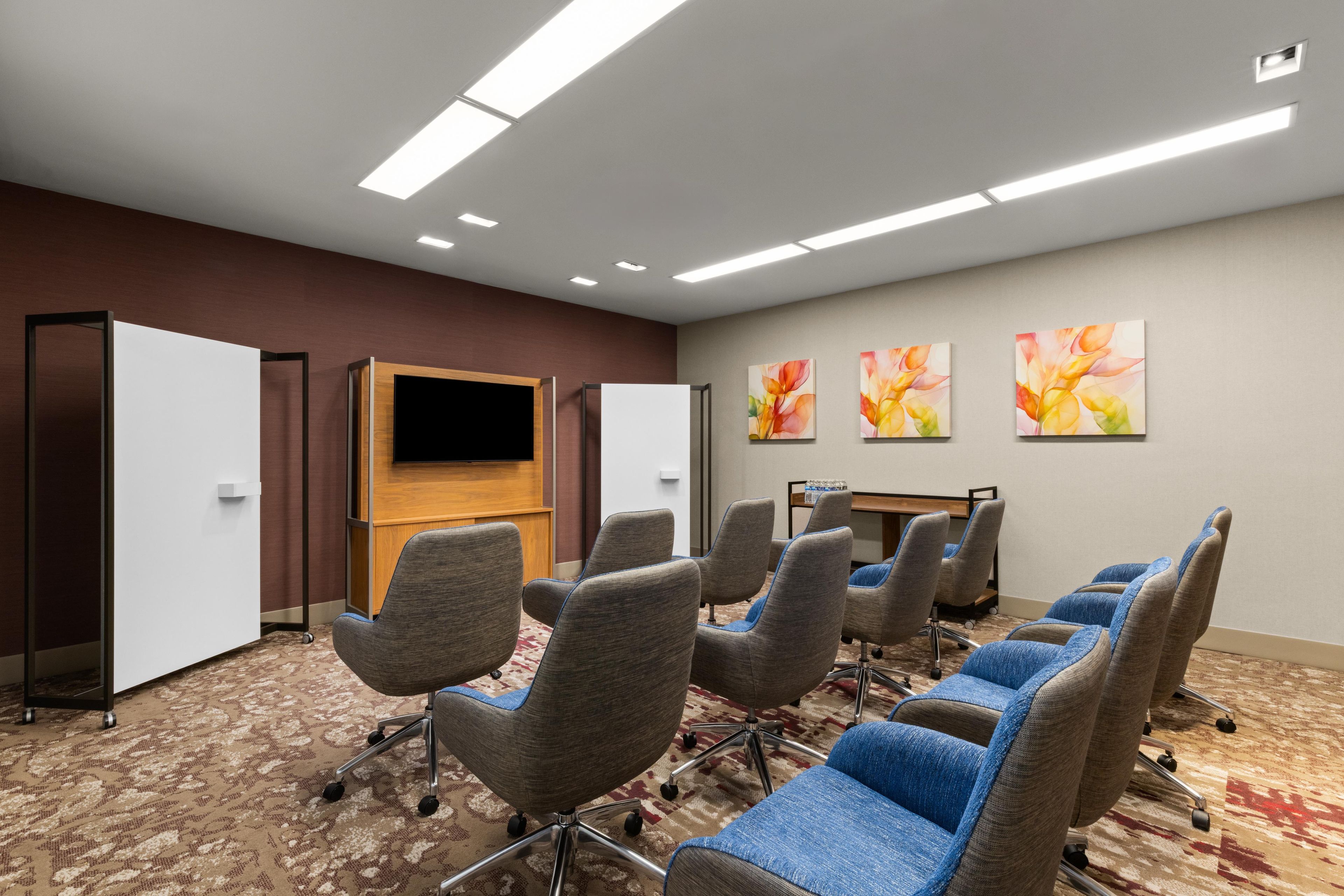 A conference room with a flat-screen TV, multiple chairs, and three floral paintings on the wall.