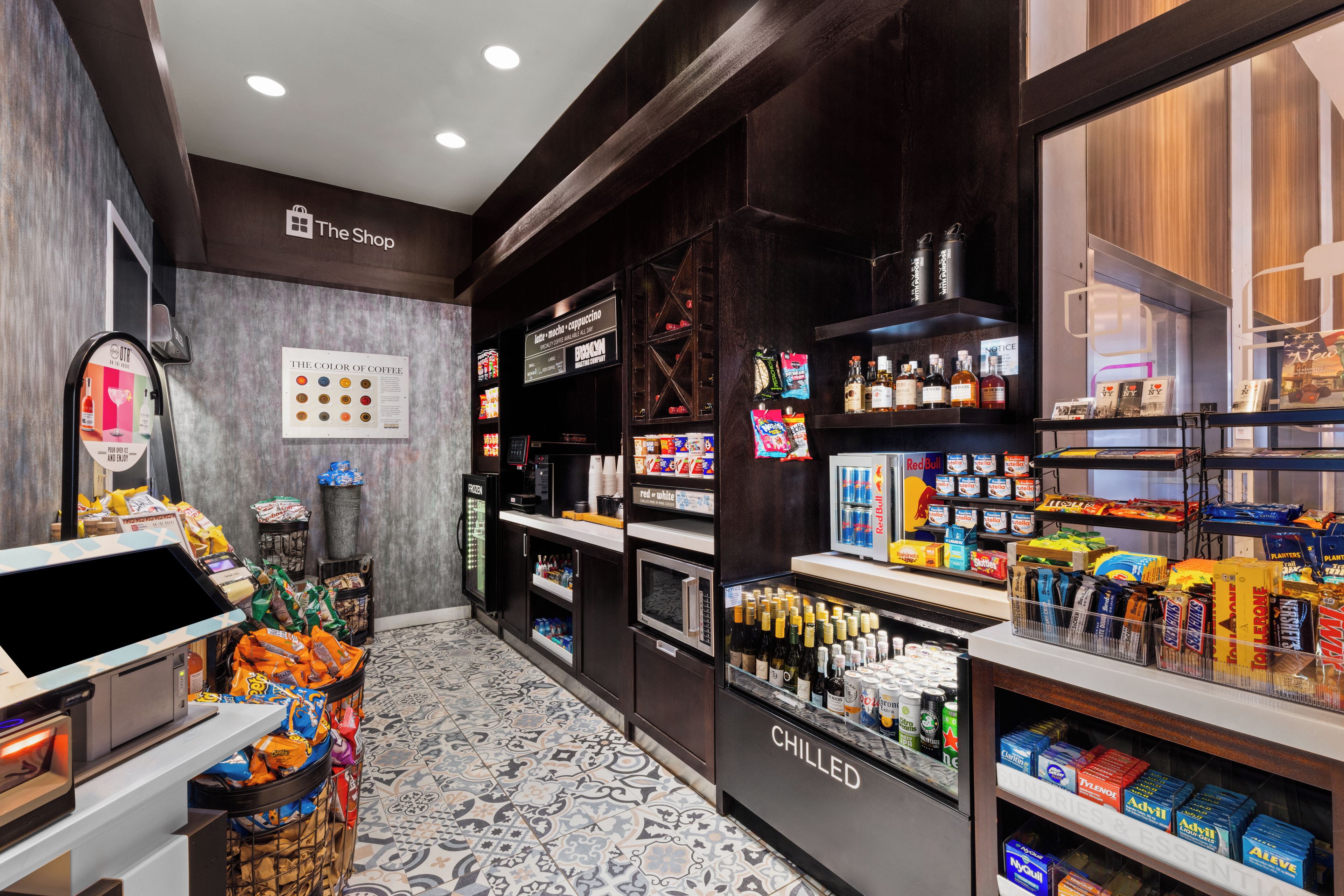 Interior de una cafetería con una máquina expendedora de café, un refrigerador con productos y estantes llenos de aperitivos y bebidas.