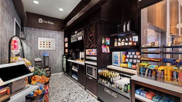 Interior de una cafetería con una máquina expendedora de café, un refrigerador con productos y estantes llenos de aperitivos y bebidas.