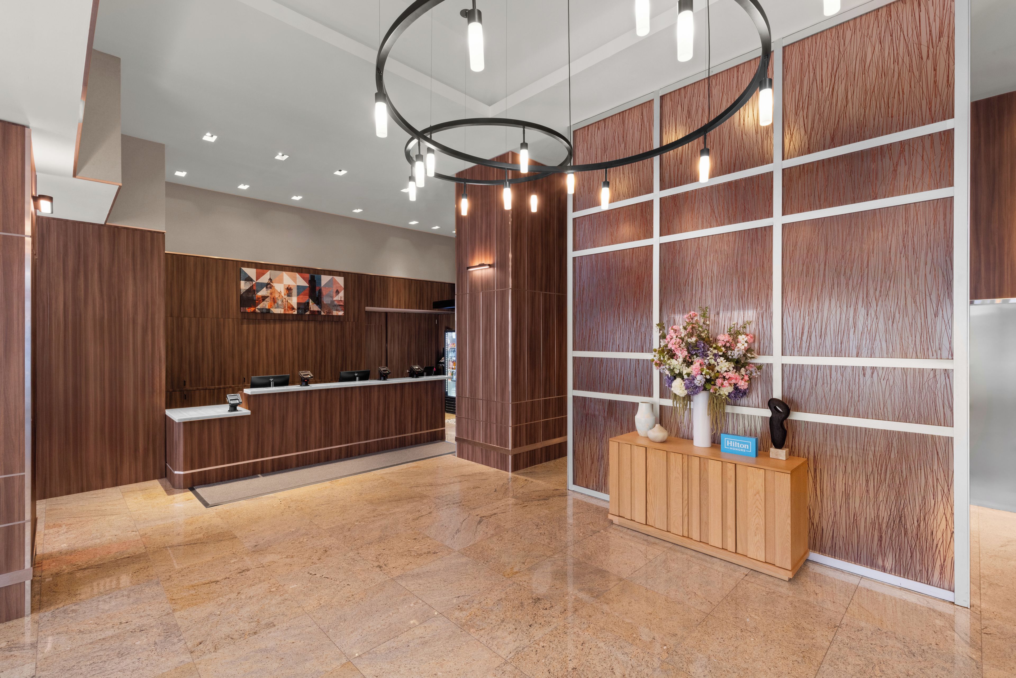 A hotel reception area with a wooden reception desk, a large floral arrangement, and a modern light fixture.