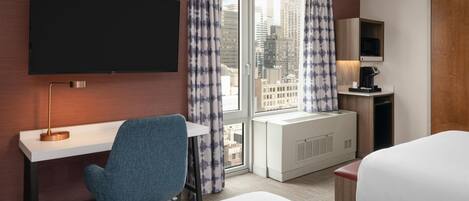 A hotel room with a bed, a desk, a chair, a TV, a coffee maker, and a view of the city.