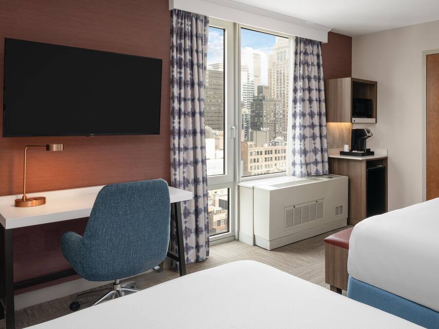 A hotel room with a bed, a desk, a chair, a TV, a coffee maker, and a view of the city.