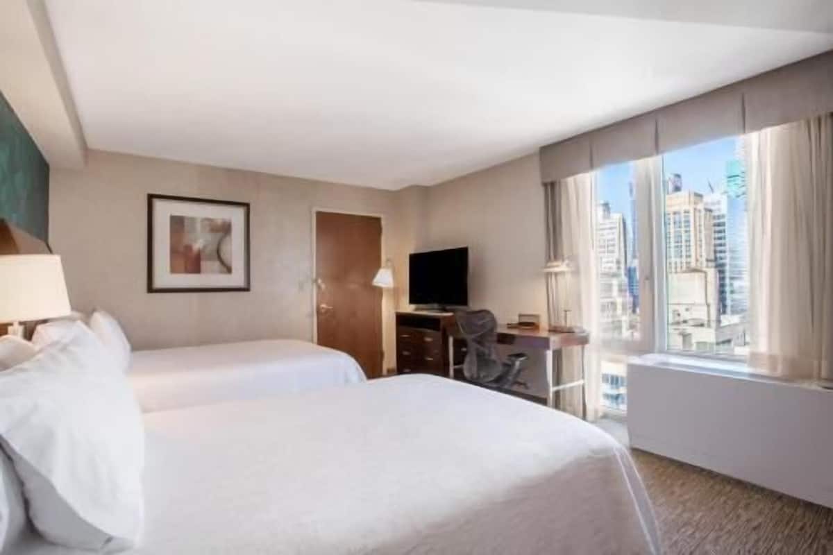 Ein Hotelzimmer mit zwei Betten, einem Schreibtisch, einem Sessel, einem Fernseher und einem großen Fenster mit Blick auf die Stadt.