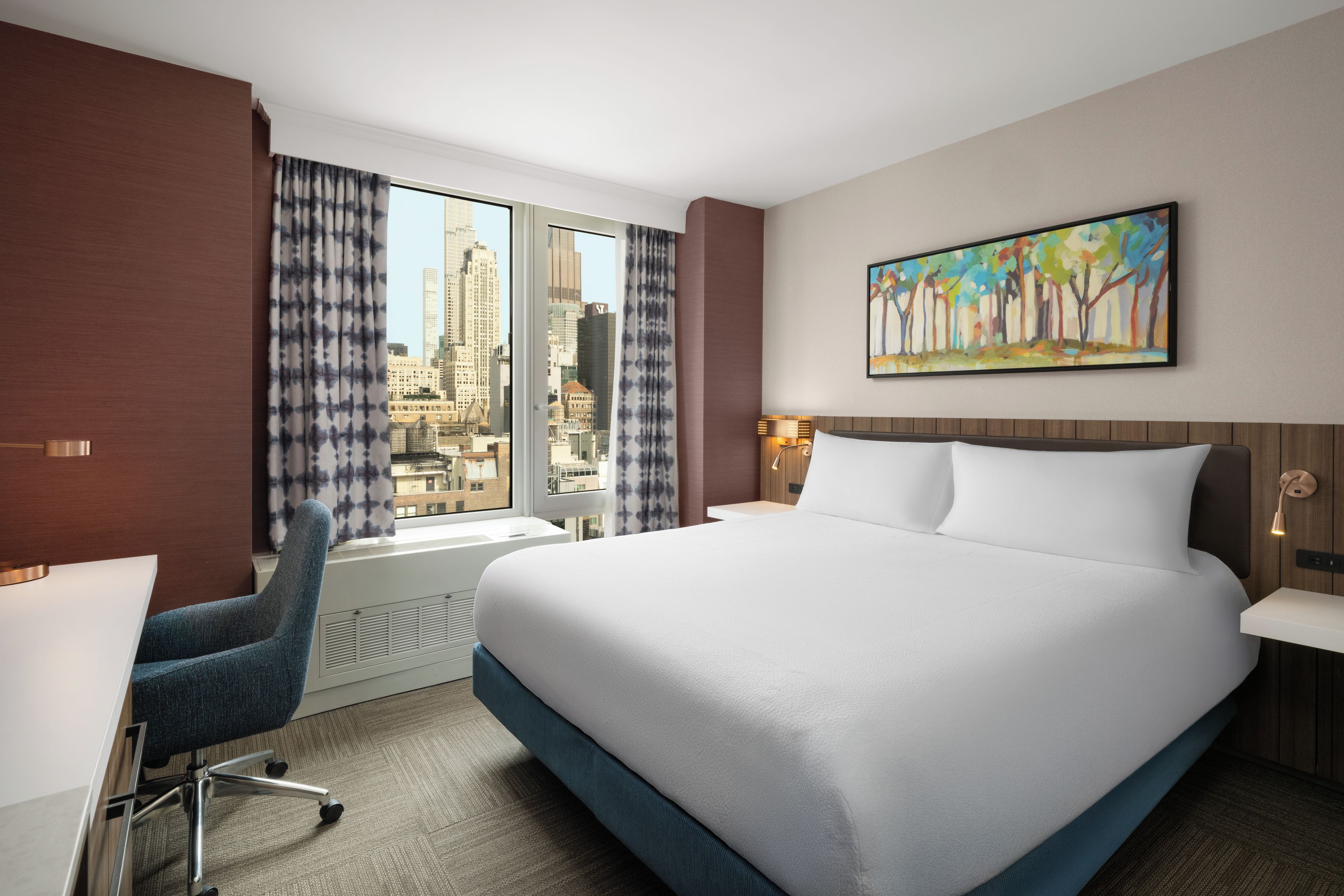 A hotel room with a large bed, a desk, a chair, a window with curtains, a painting on the wall, and a city view.