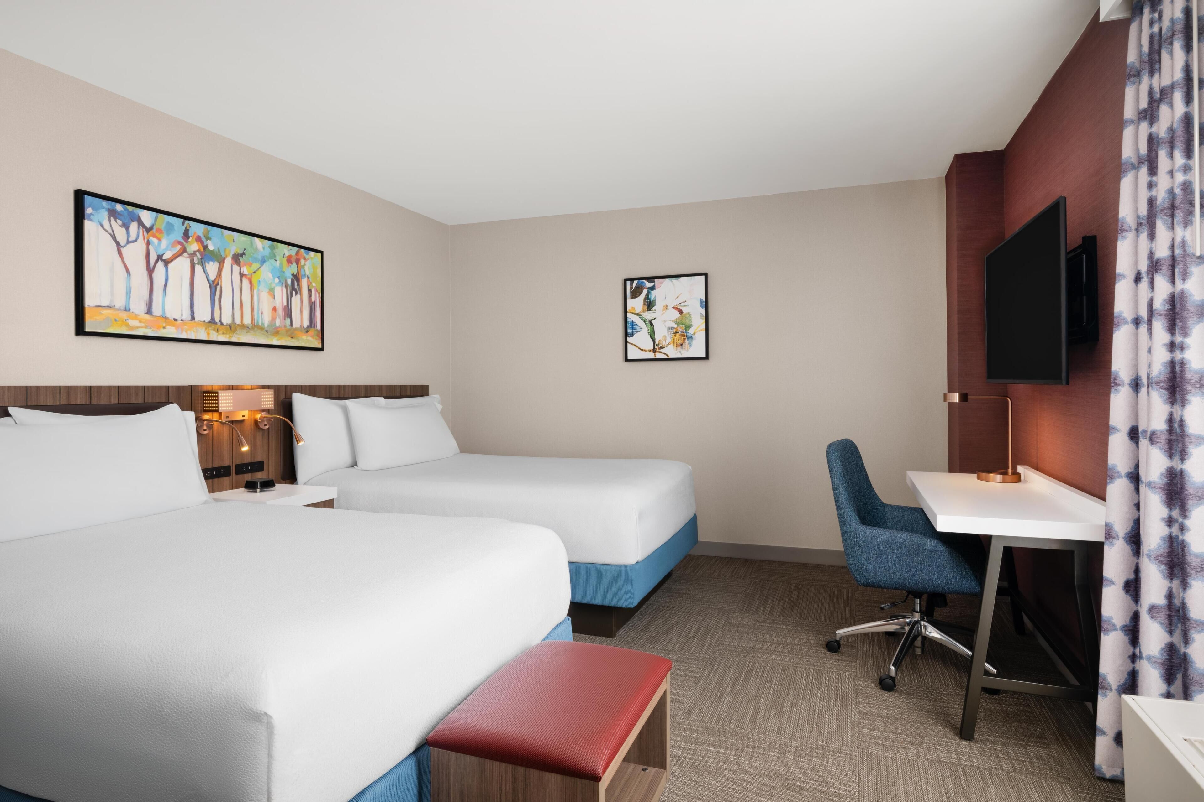 A hotel room with two beds, a desk, a chair, a TV, and artwork on the walls.