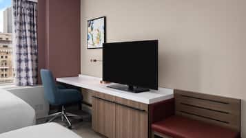 Une chambre d’hôtel avec un lit, un bureau avec une télévision, une chaise, un banc rouge et une fenêtre avec des rideaux.