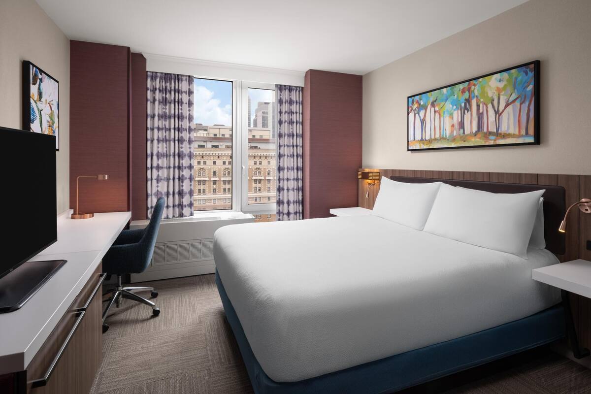 Une chambre d’hôtel avec un lit, un bureau, une chaise et une grande fenêtre avec des rideaux.