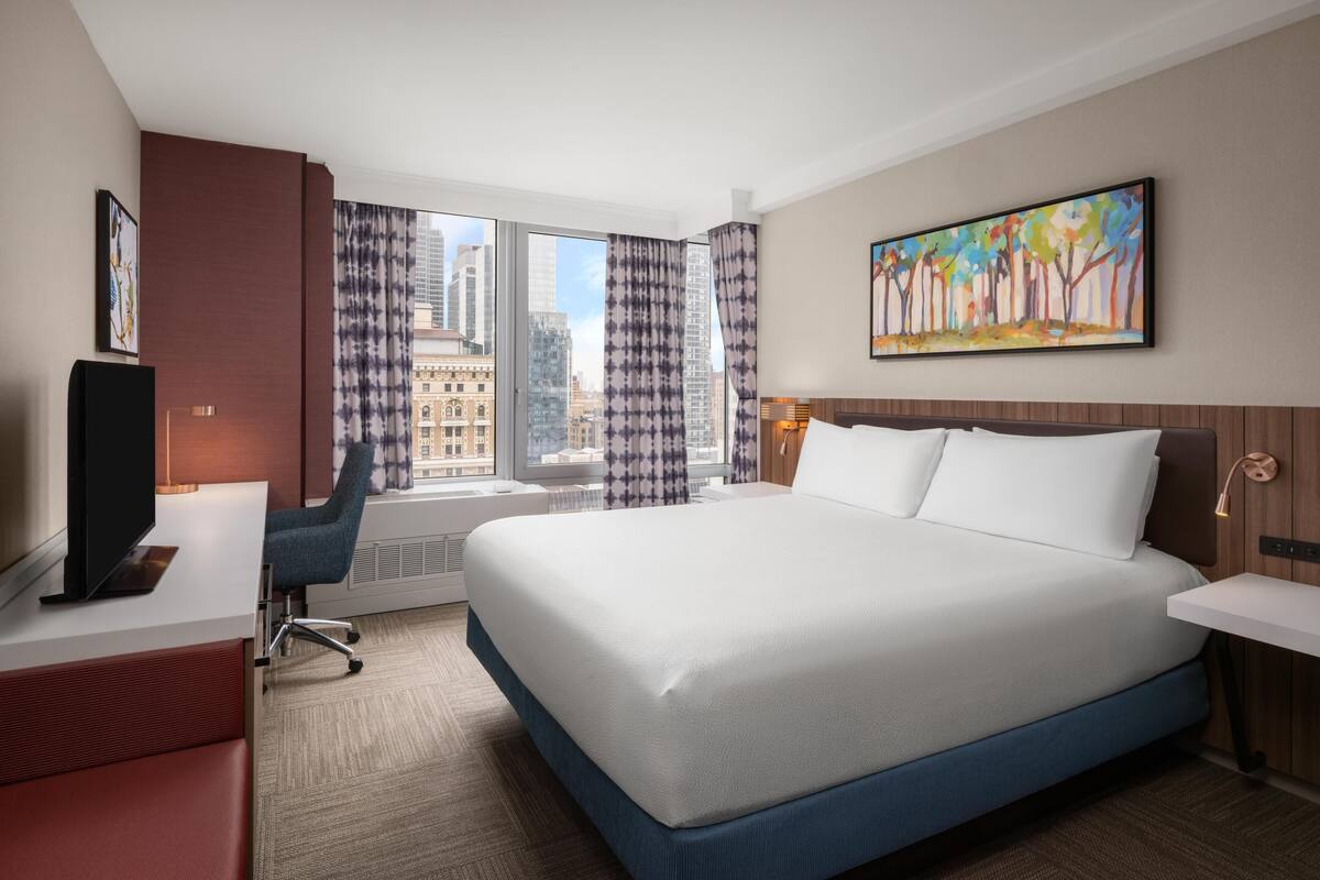 Une chambre d’hôtel avec un grand lit, un bureau avec une chaise, une télévision et une fenêtre avec des rideaux.