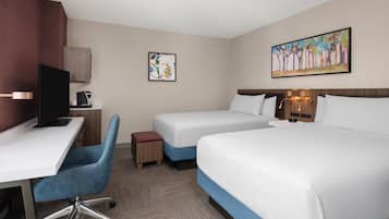 A hotel room with a desk, two beds, a chair, a small table, and artwork on the walls.