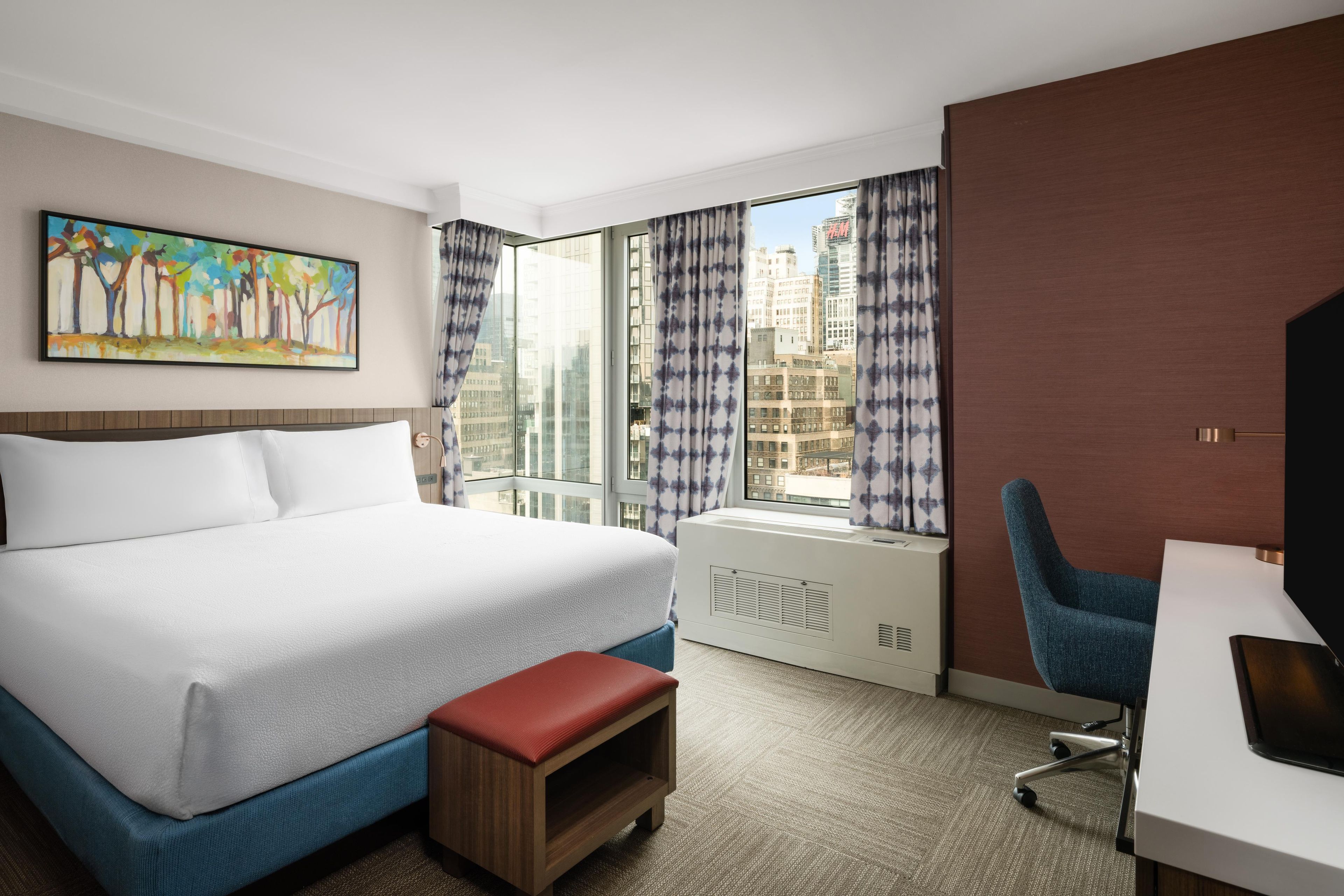 A hotel room with a bed, a desk, a chair, a window with curtains, and a painting on the wall.