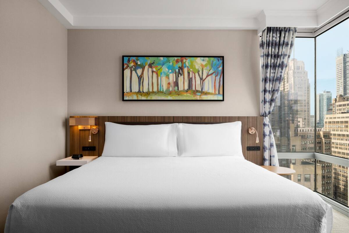 Une chambre d’hôtel avec un grand lit, une table de chevet avec une lampe, un tableau au mur et une vue sur la ville par la fenêtre.