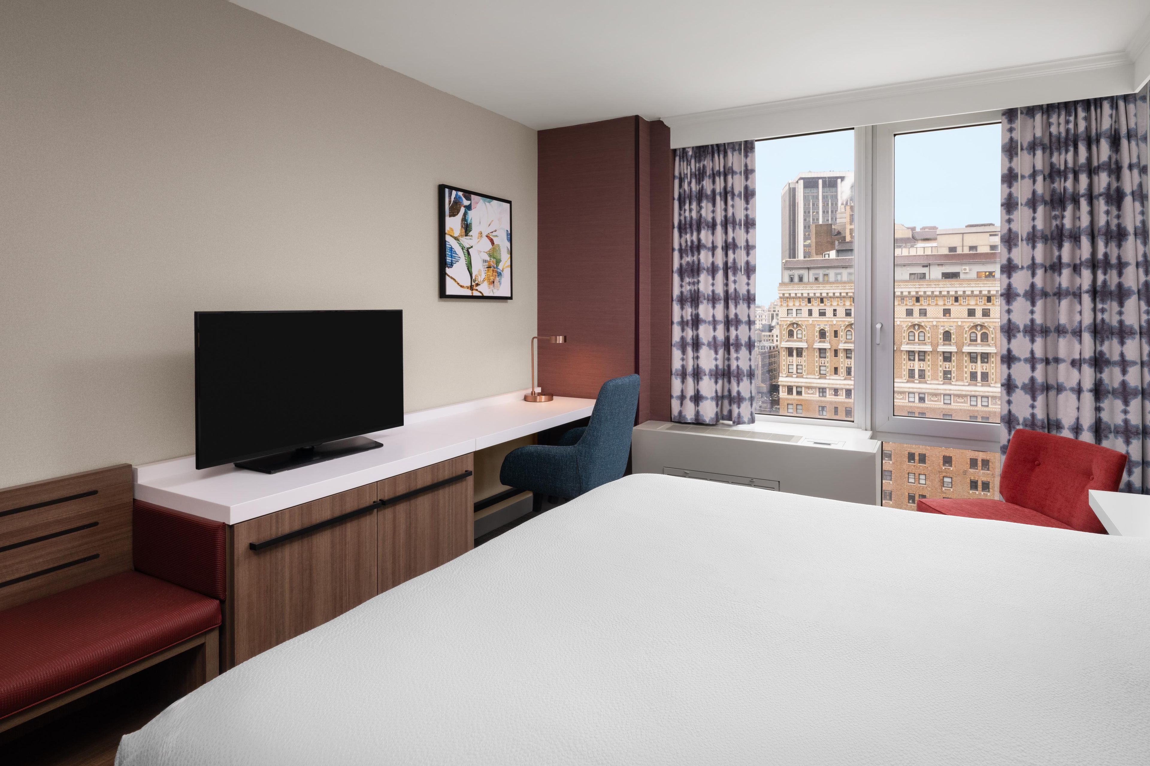A hotel room with a bed, a desk with a television, a chair, and a window with curtains.