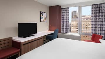 Quarto de hotel com cama, escrivaninha com televisão, uma cadeira e janela com cortinas.