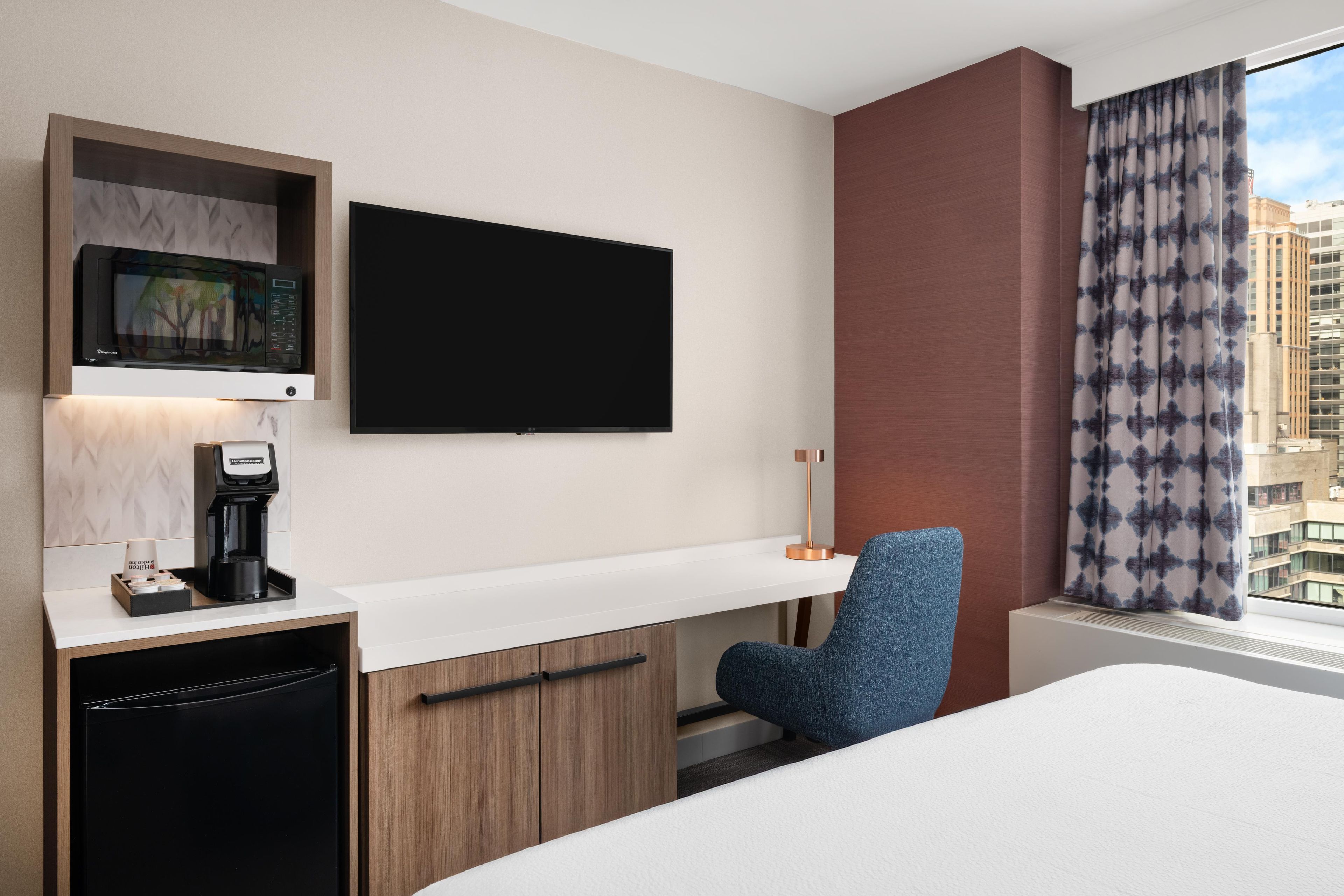A hotel room with a bed, a desk with a coffee maker, a TV, and a window with curtains.