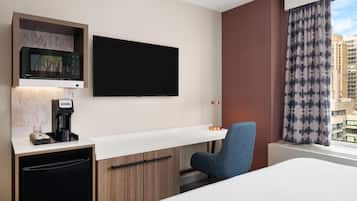 A hotel room with a bed, a desk with a coffee maker, a TV, and a window with curtains.