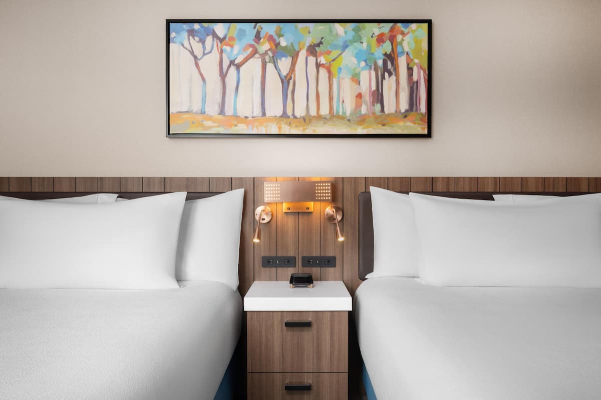 Une chambre d’hôtel avec deux lits, une tête de lit en bois, une table de chevet avec une lampe et un tableau au mur.