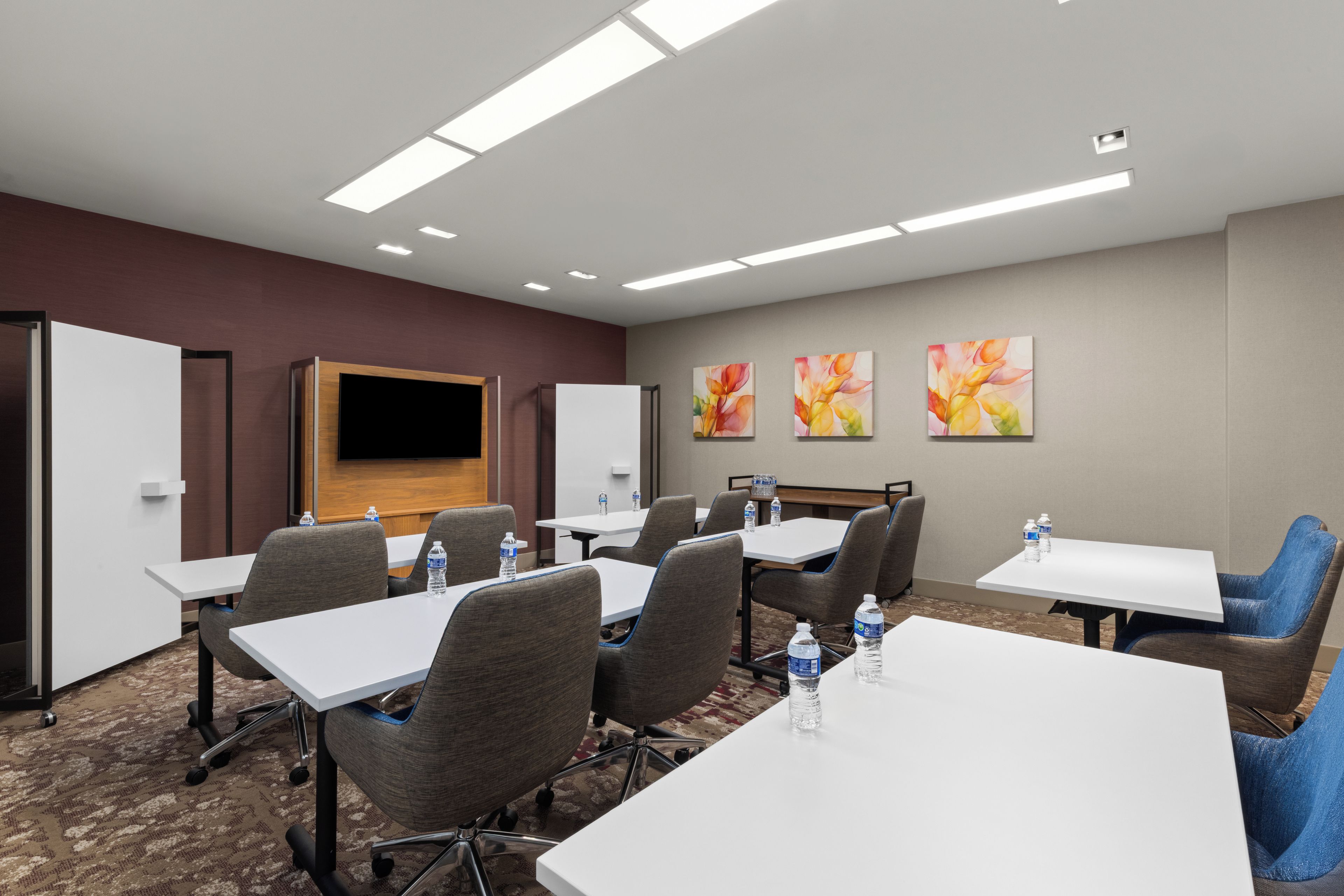 A conference room with white tables, grey chairs, and a flat-screen TV mounted on the wall.