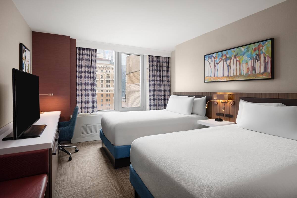 Une chambre d’hôtel avec deux lits, un bureau doté d’un téléviseur à écran plat, une fenêtre avec des rideaux et un tableau accroché au mur.