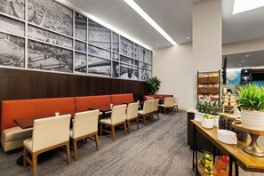 Eine Hotellobby mit einem roten Sofa, Esstischen, Stühlen und einer Wand mit schwarz-weißen Stadtansichten sowie einem Fernseher an der Wand.