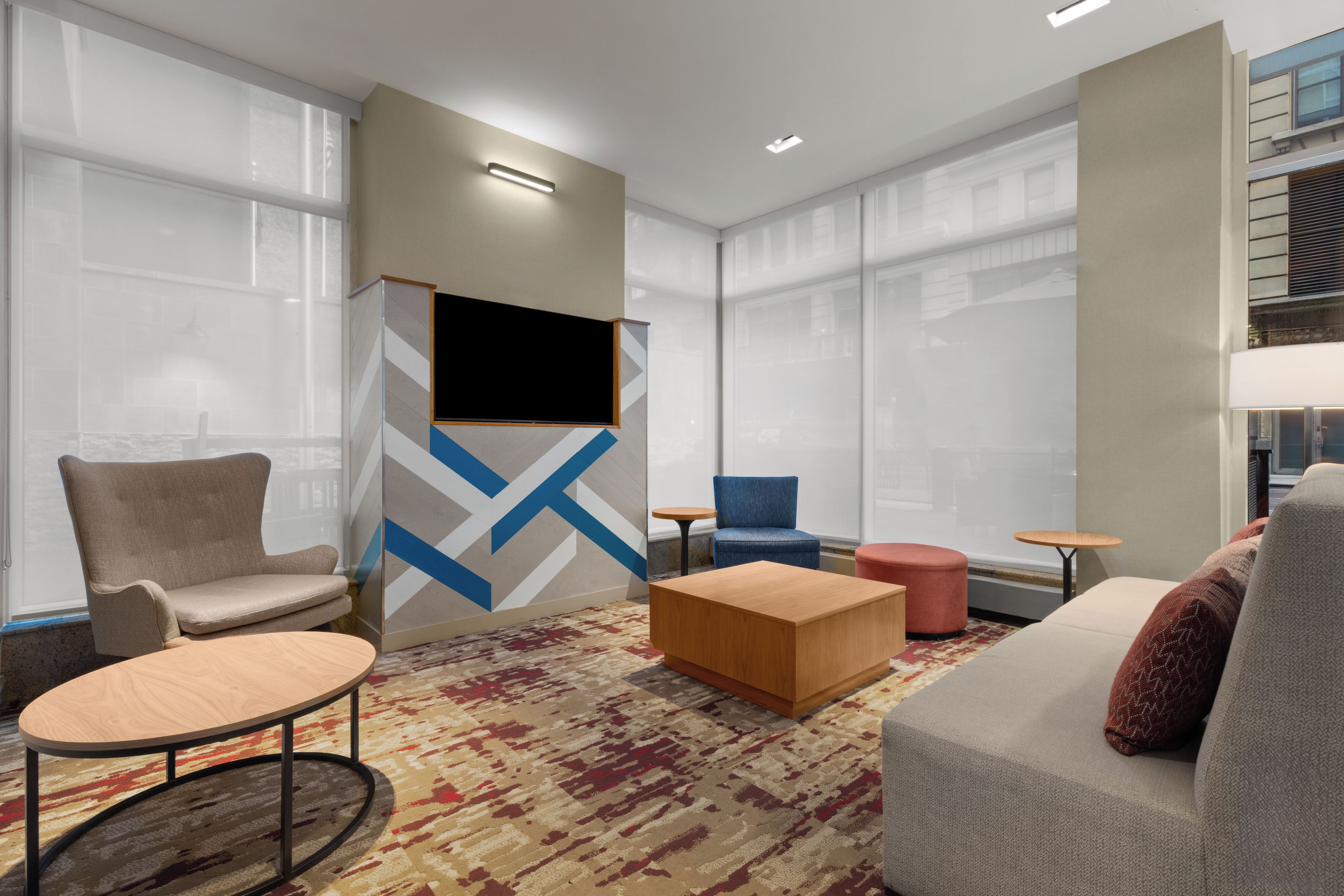 A modern hotel lobby with a seating area, a flat-screen TV mounted on a wall, and large windows.