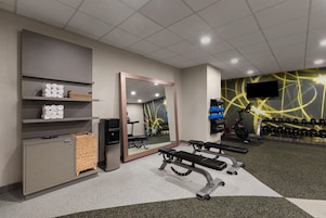 A gym with exercise equipment, a mirror, shelves with towels, and a wall-mounted TV.