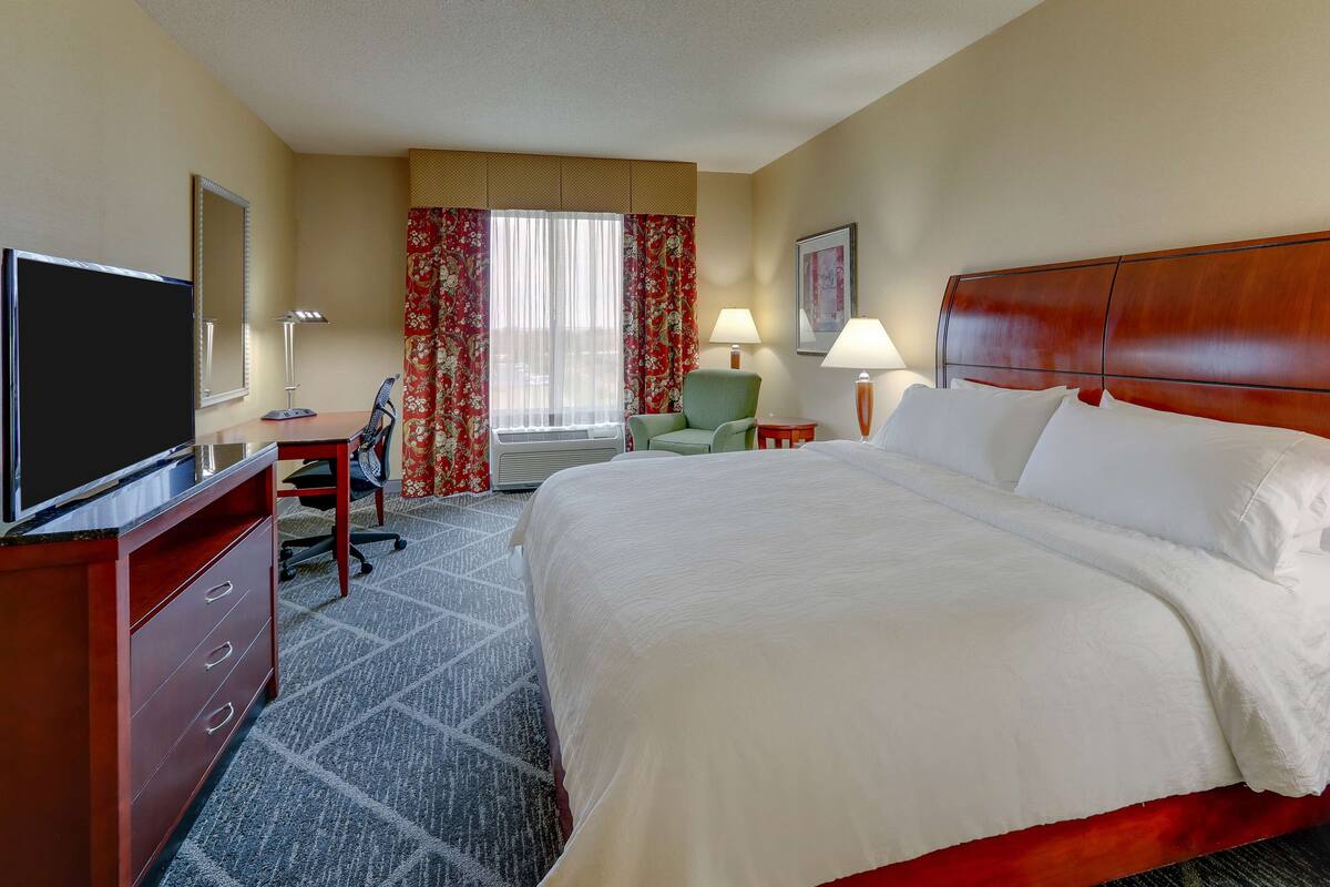 Une chambre d’hôtel avec un grand lit, un bureau avec une télévision, une chaise, une lampe et une fenêtre avec des rideaux.
