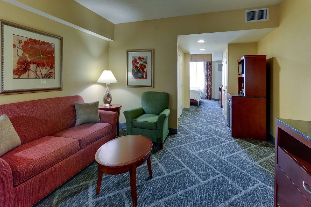 Une chambre d’hôtel avec un canapé rouge, un fauteuil vert et une petite table en bois. Deux tableaux encadrés représentant des motifs floraux ornent les murs, ainsi qu’un meuble en bois.