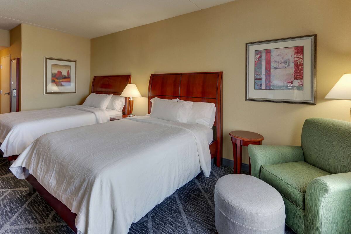 Une chambre d’hôtel avec deux lits, un fauteuil vert, une petite table ronde et deux lampes fixées au mur.