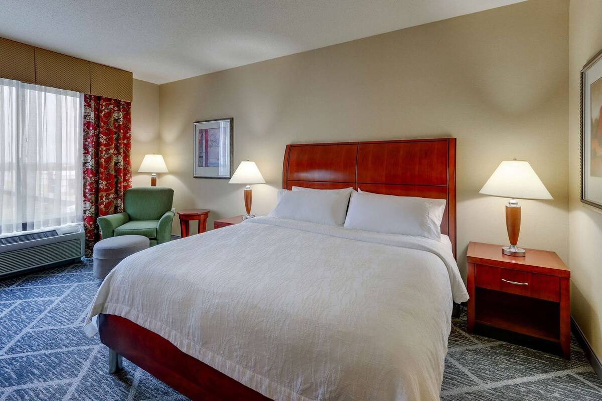 Une chambre d’hôtel avec un grand lit, deux tables de chevet avec des lampes, un fauteuil vert, une chaise rouge, un tableau encadré au mur et une fenêtre avec des rideaux.