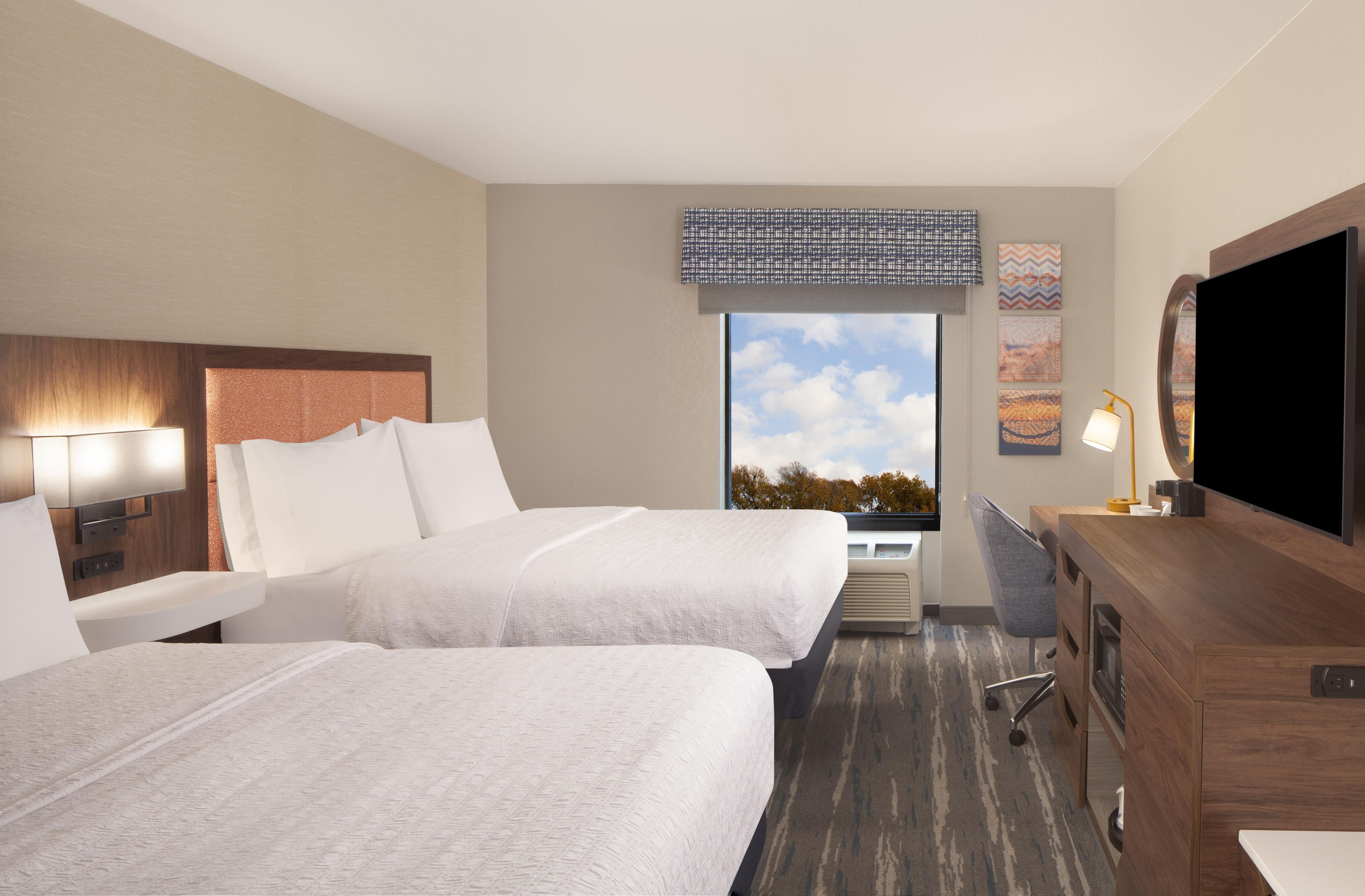 A hotel room with two beds, a desk, a television, and a window with a view of trees.