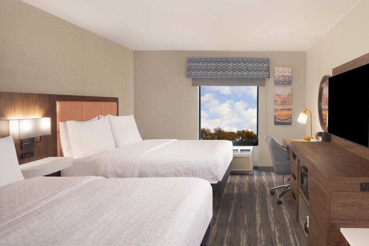 A hotel room with two beds, a desk, a television, and a window with a view of the sky.
