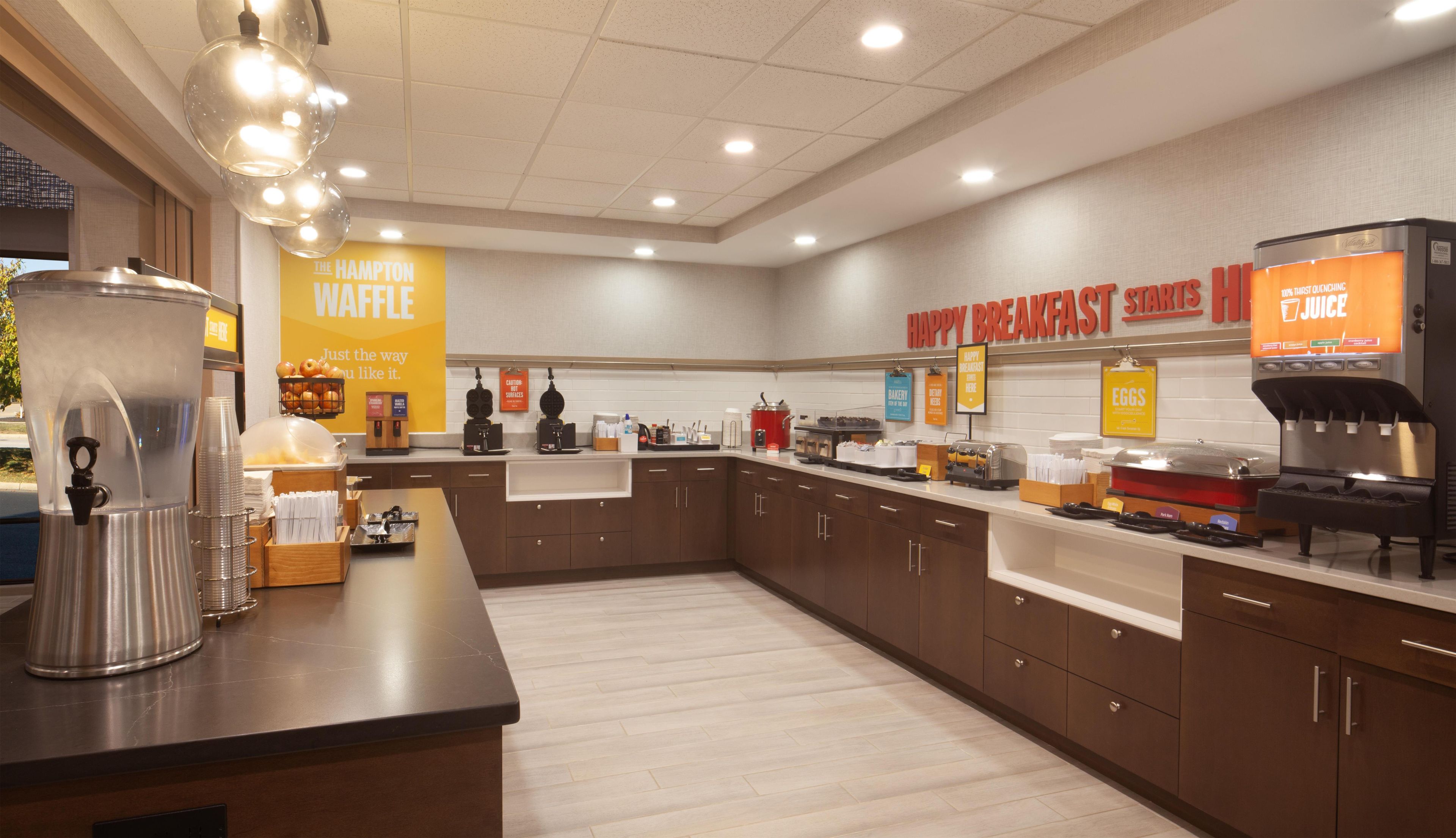 A breakfast area with a waffle station, juice dispenser, and various breakfast items.