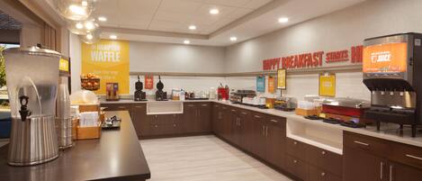 A breakfast area with a waffle station, juice dispenser, and various breakfast items.