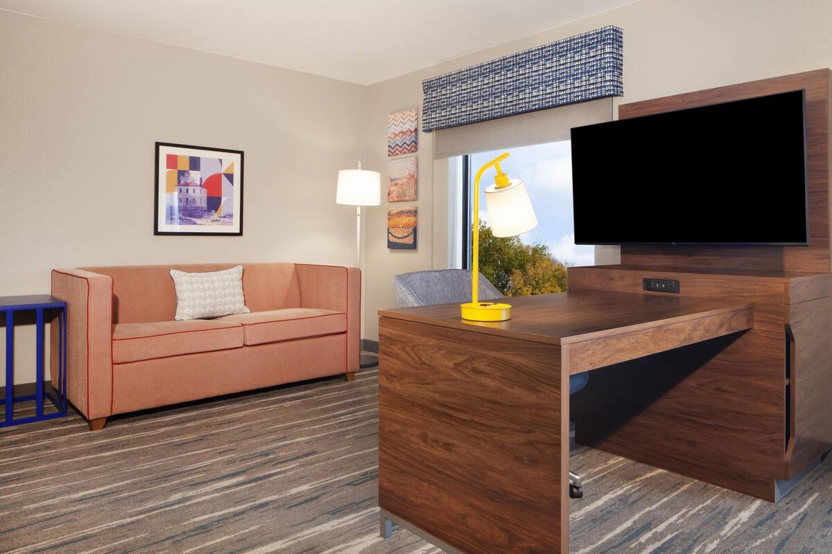 A hotel room with a wooden desk, a flat-screen TV, a sofa, and a patterned carpet.
