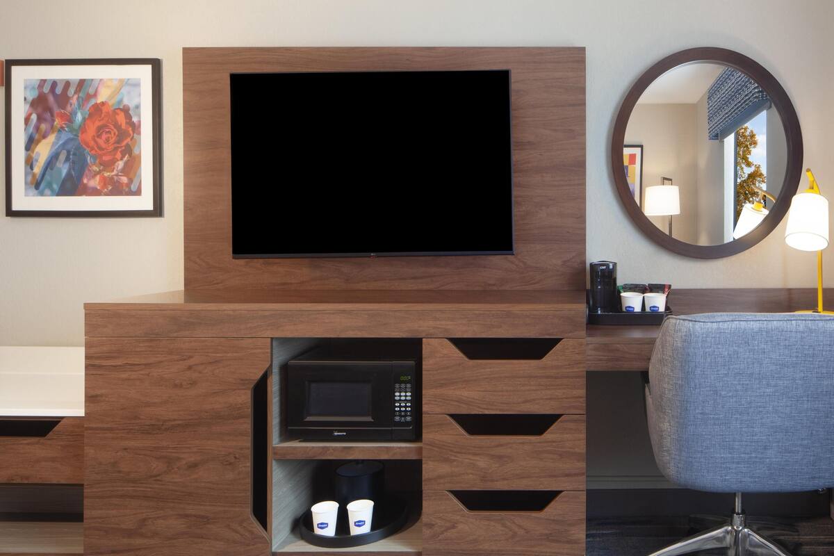 A modern hotel room with a flat-screen TV mounted on a wooden cabinet, a microwave below it, and a seating area with a chair and a round mirror.
