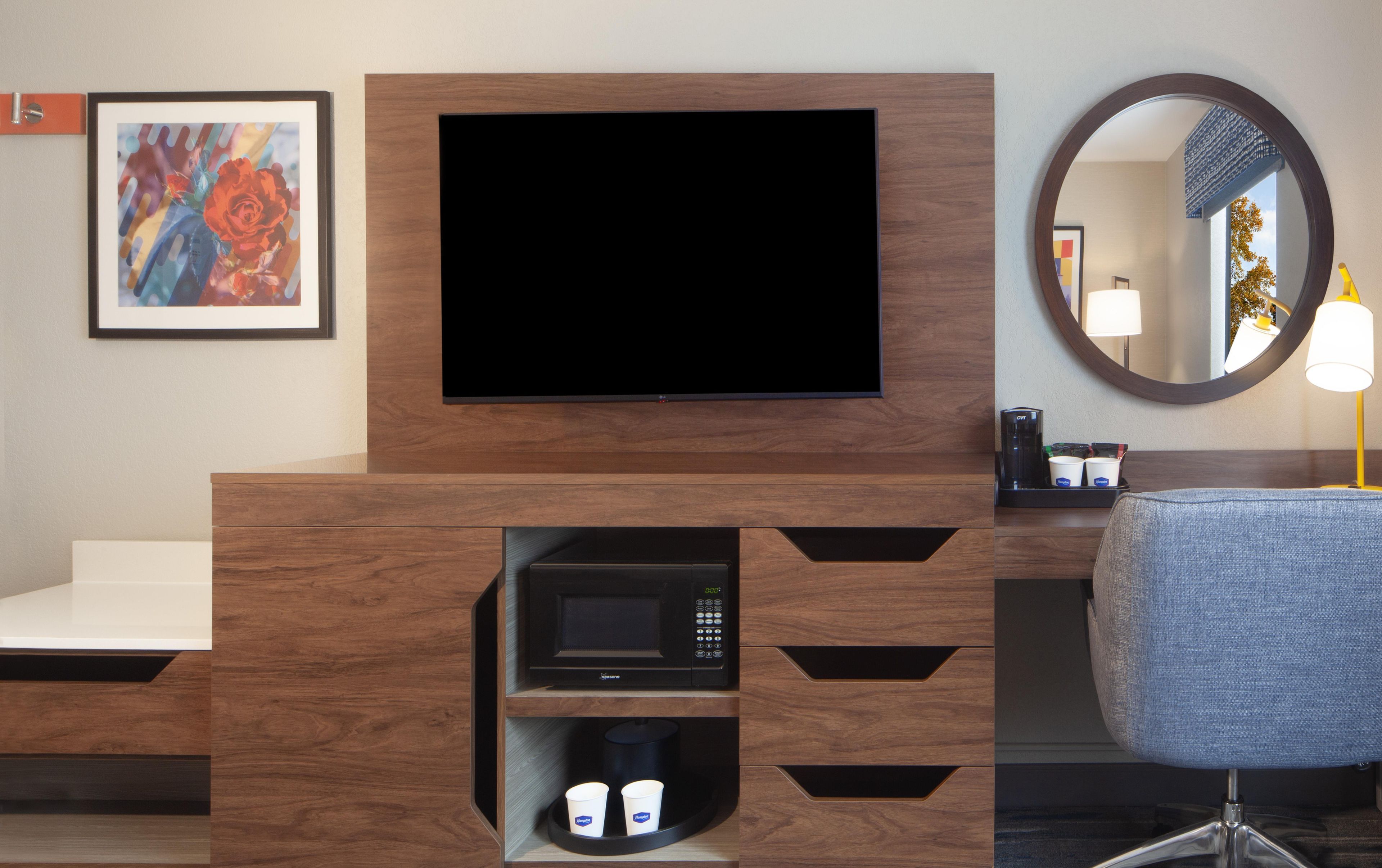 Ein Hotelzimmer mit einem Flachbildfernseher, der an einem Holzschrank befestigt ist, einer Mikrowelle darunter und einer Sitzecke mit einem Sessel und einem runden Spiegel.