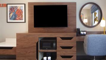 A hotel room with a flat-screen TV mounted on a wooden cabinet, a microwave below it, and a seating area with a chair and a round mirror.