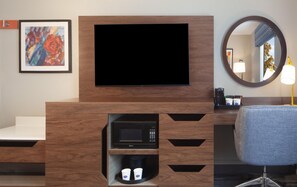 Habitación de hotel con televisor de pantalla plana montado en un mueble de madera, microondas debajo y una zona de estar con una silla y un espejo redondo.