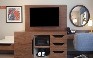A hotel room with a flat-screen TV mounted on a wooden cabinet, a microwave below it, and a seating area with a chair and a round mirror.