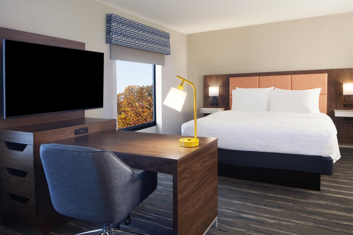 Habitación de hotel con una cama grande, un escritorio con silla, un televisor y ventana con vista a árboles otoñales.