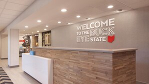 A reception area with a wooden reception desk, a wall sign reading "WELCOME TO THE BUCK EYE STATE," and a coffee machine in the background. - Hampton Inn & Suites Cleveland Mentor (Mentor)
