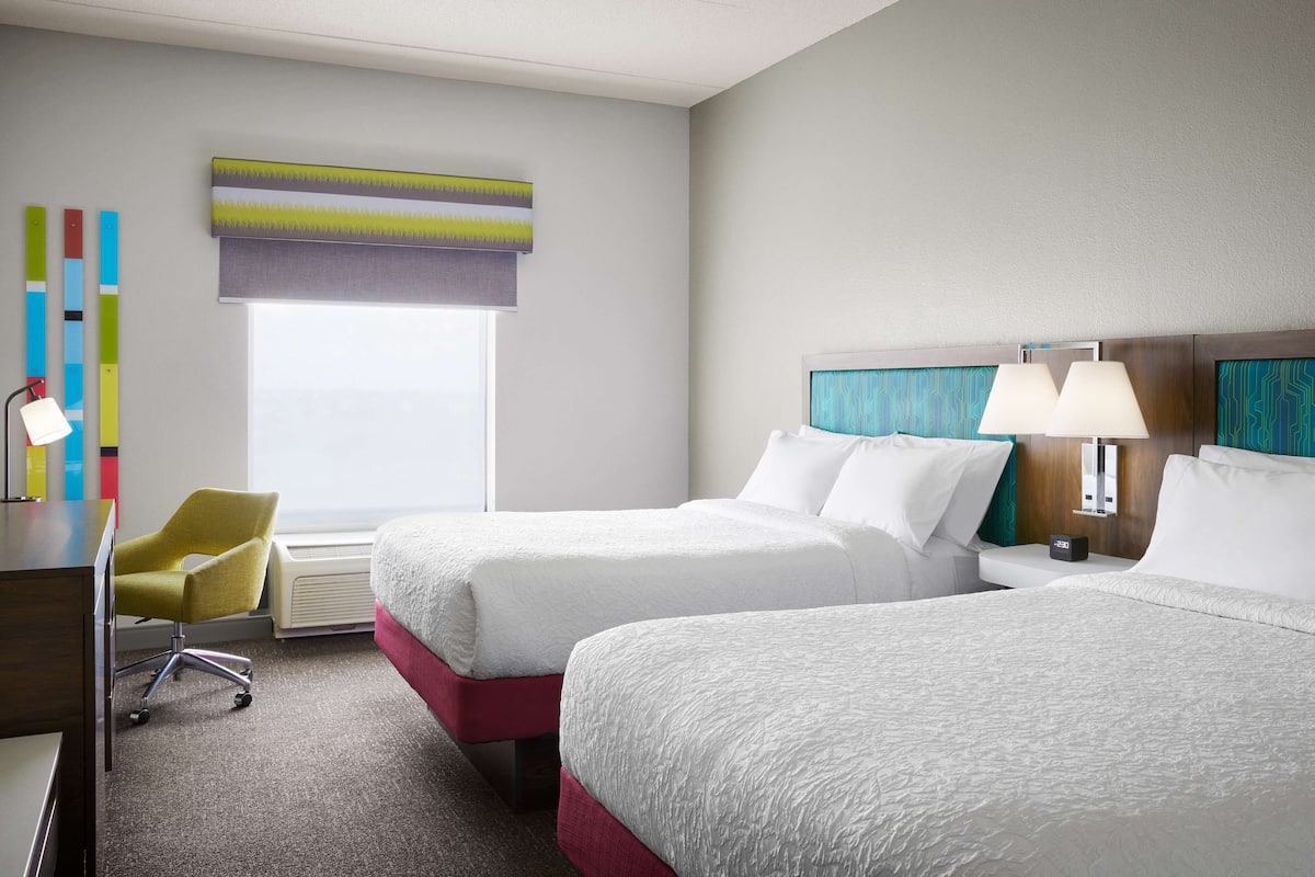 A hotel room with two beds, a desk, a chair, and a window with blinds.