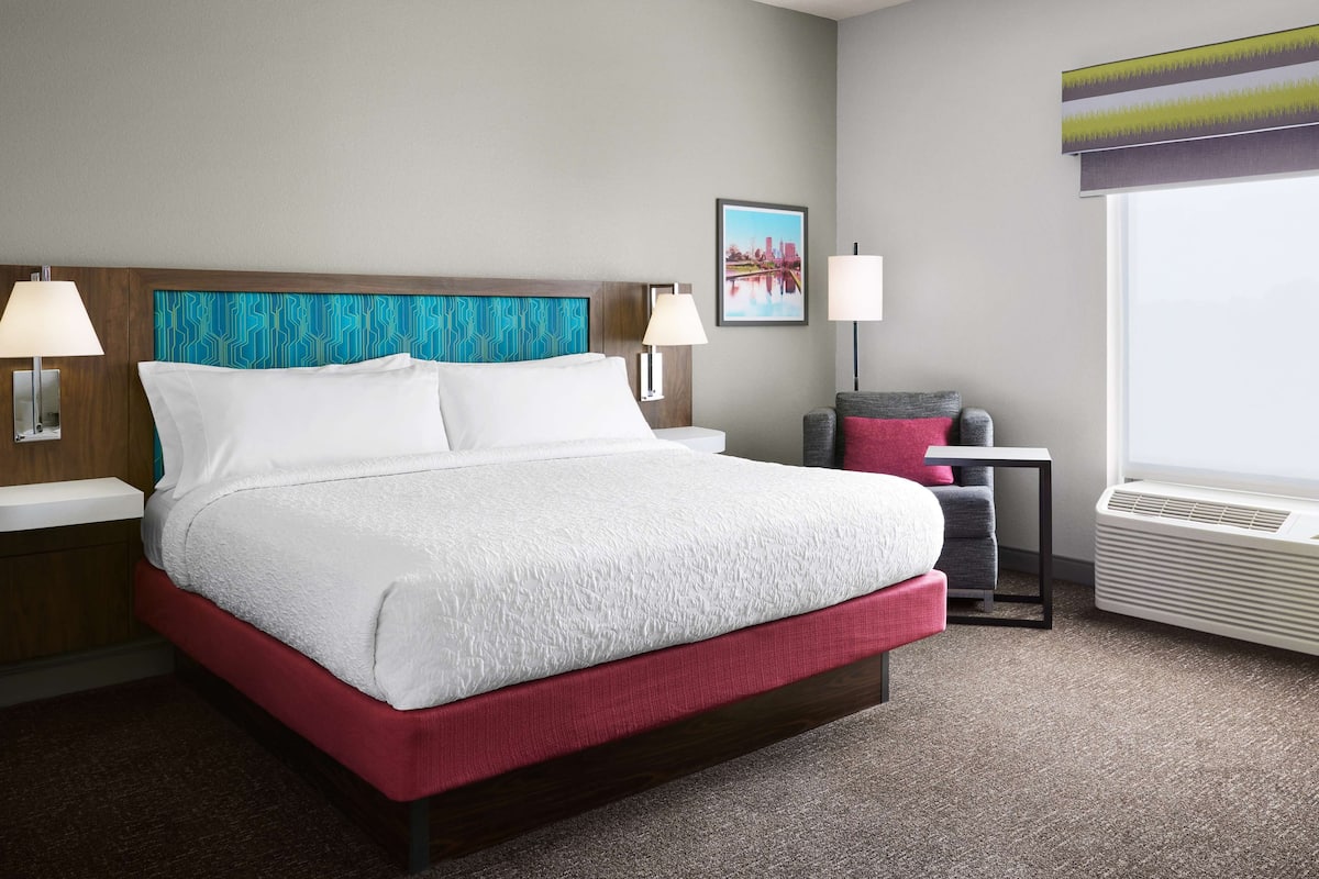 A hotel room with a large bed, two bedside tables, a chair, a window with blinds, and a wall-mounted air conditioner.