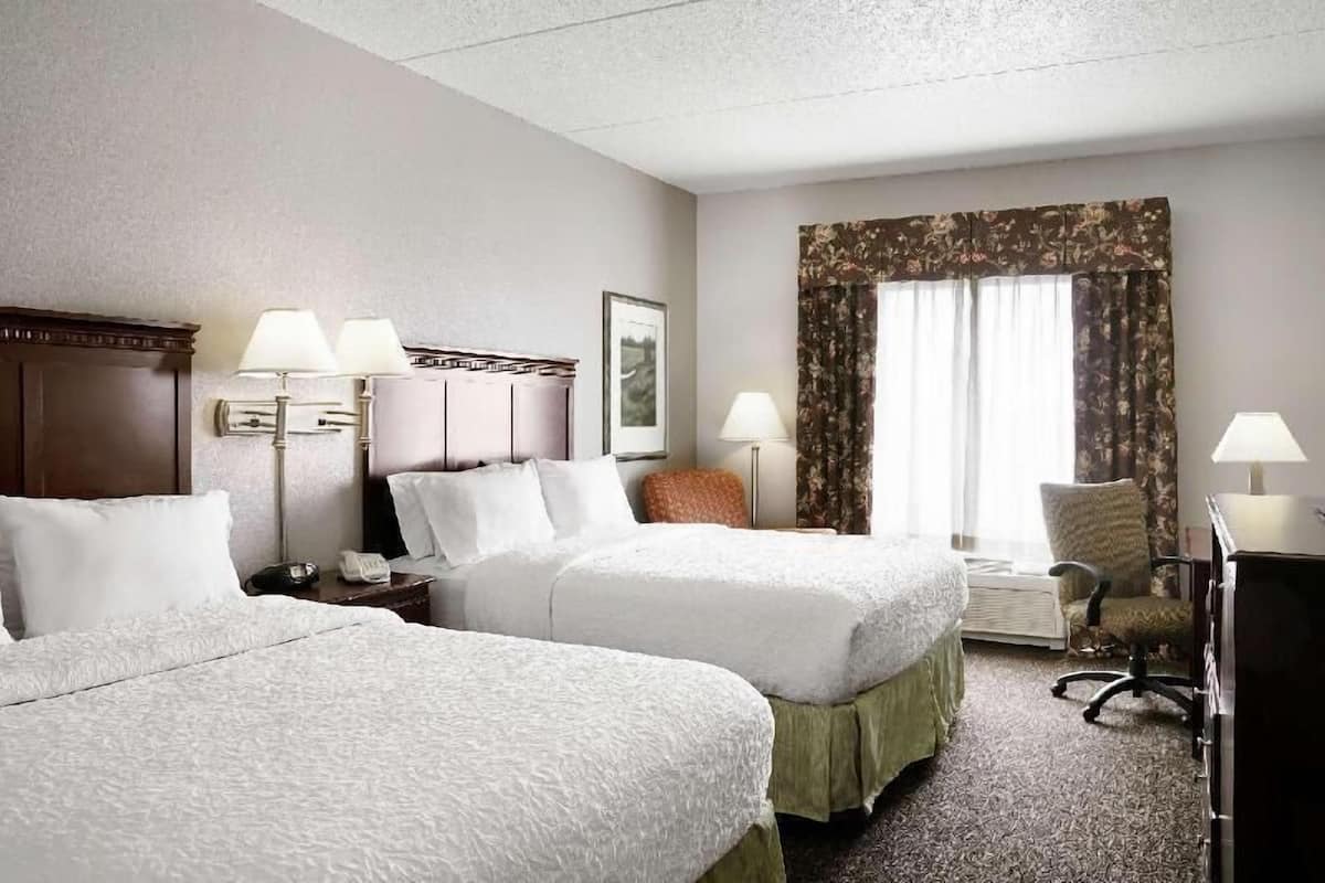 A hotel room with two beds, a desk, a chair, and a window with curtains.