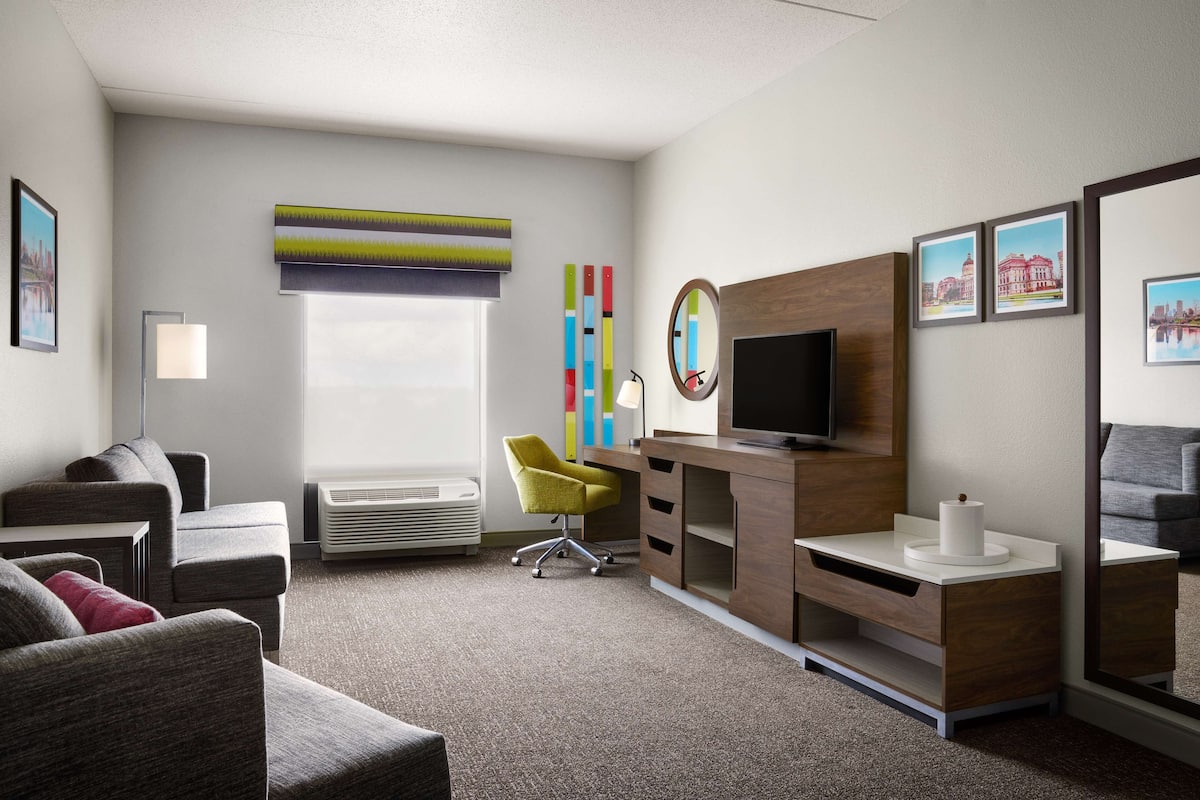 A modern hotel room with a grey sofa set, a yellow chair, a wooden desk with a flat-screen TV, and a large window with a view of a building.