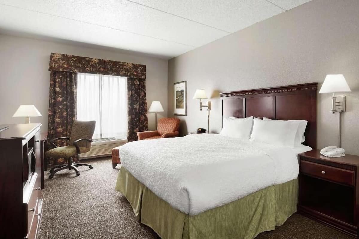 A hotel room with a large bed, a desk, a chair, two bedside lamps, and a window with curtains.