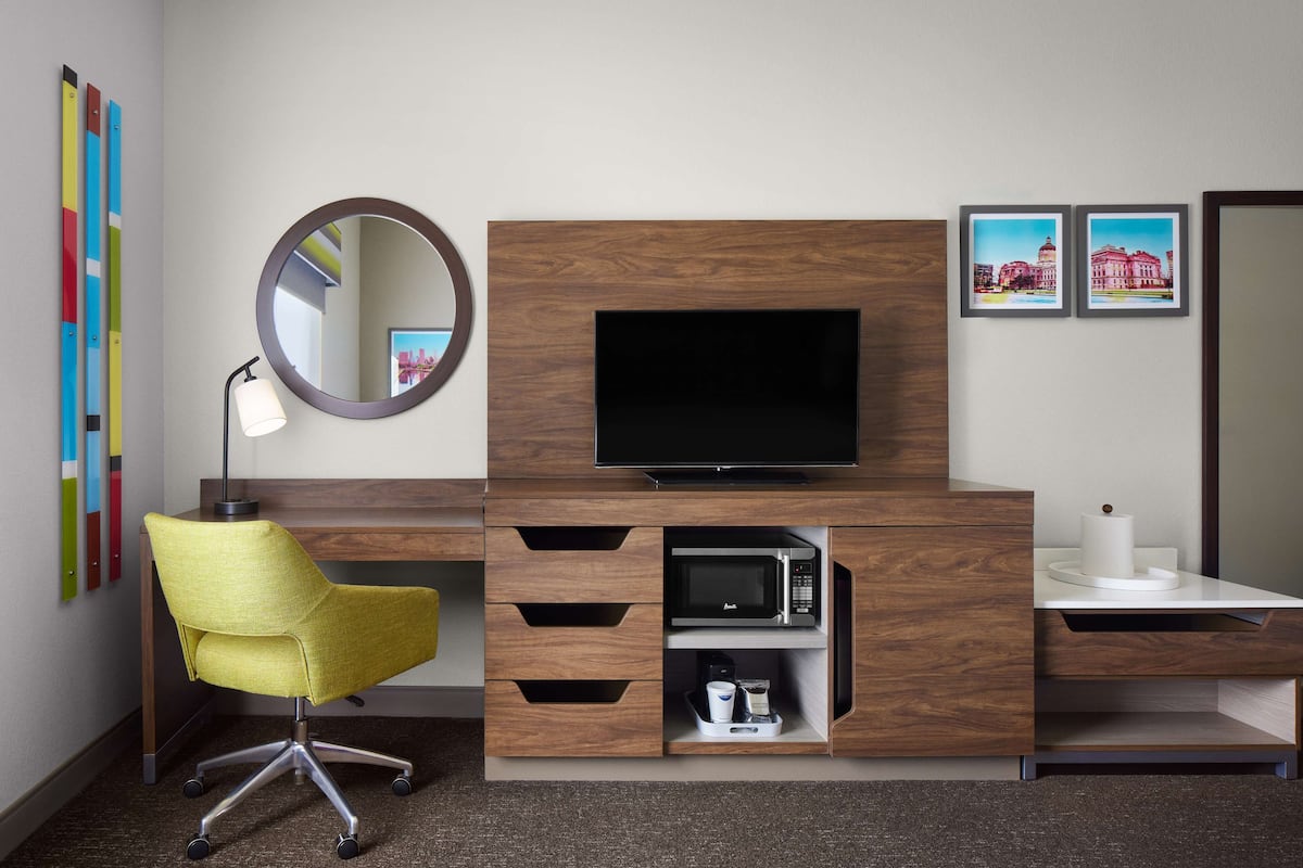A hotel room with a wooden desk, a television, a yellow chair, a mirror, and two framed pictures on the wall.