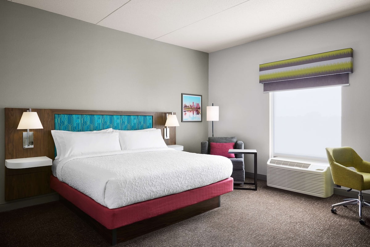 A hotel room with a large bed, two bedside tables with lamps, a window with a view, a chair, and a heater.