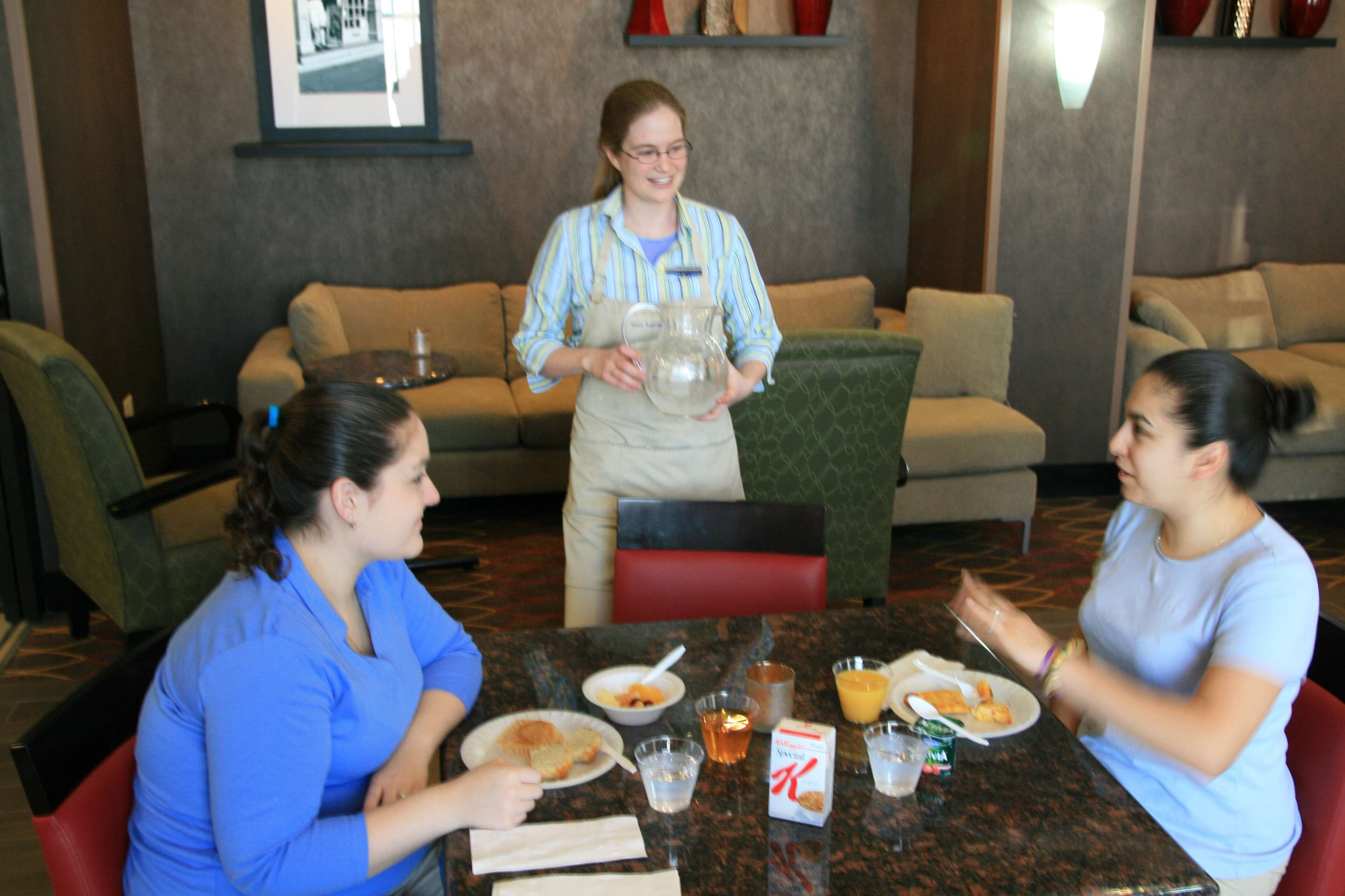 A dining area with a table set for three people, a waiter holding a pitcher, and a couch in the background.